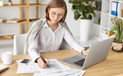 Serious woman wears formal white blouse, works in office, fills in business documents, surrounded with laptop and smart phone. Female boss prepares necessary papers for presenting on meeting